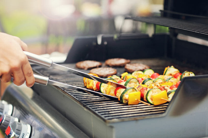 A person uses tongs to grill vegetable and meat skewers next to several hamburger patties on an outdoor barbecue grill.