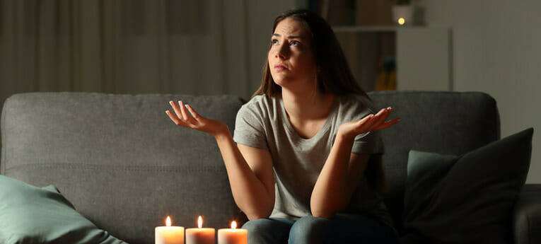 A woman sits on a sofa in a dimly lit room with three lit candles in front of her, looking up and shrugging her shoulders.