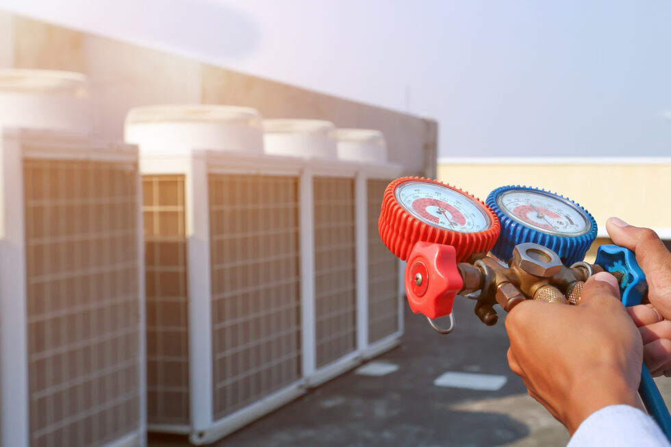 Person holding HVAC manifold gauge in front of multiple outdoor air conditioning units on a rooftop, checking system pressure levels.