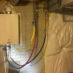 %Imagetitle% | Sal'S Heating &Amp; Cooling, Plumbing &Amp; Sewer Tankless water heater installed on a basement wall with red, blue, and yellow pipes connected and insulation on adjacent surfaces.