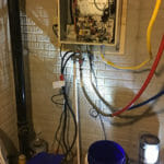%Imagetitle% | Sal'S Heating &Amp; Cooling, Plumbing &Amp; Sewer An open electrical panel with exposed wires is mounted on a brick wall. Various pipes and a blue bucket are visible below the panel.