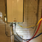 %Imagetitle% | Sal'S Heating &Amp; Cooling, Plumbing &Amp; Sewer A tankless water heater mounted on a brick wall with various colored pipes connected to it.