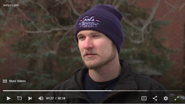 A man wearing a navy blue beanie and a black jacket stands outdoors in front of trees, speaking during an interview. Video player controls are visible at the bottom.