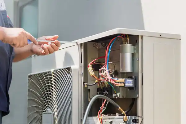 A person uses a screwdriver to repair or maintain an outdoor air conditioning unit with exposed wires and components.