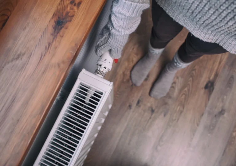 Person wearing gray gloves, sweater, and socks adjusts the temperature on a white radiator next to a wooden surface on a hardwood floor.