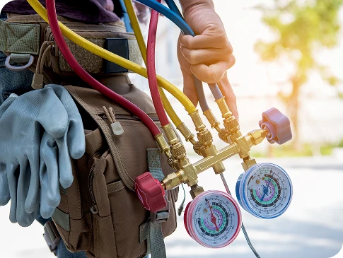 A person holds HVAC manifold gauges and hoses, with work gloves and a brown tool bag visible on their side.