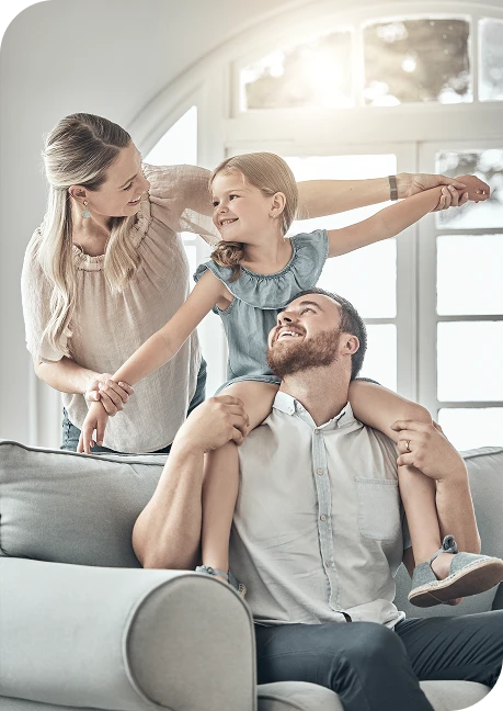 A man sits on a couch with a young girl on his shoulders while a woman stands nearby; all three are smiling and interacting in a bright living room.