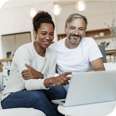 Two people sitting on a couch, smiling and looking at a laptop screen together in a bright living room.