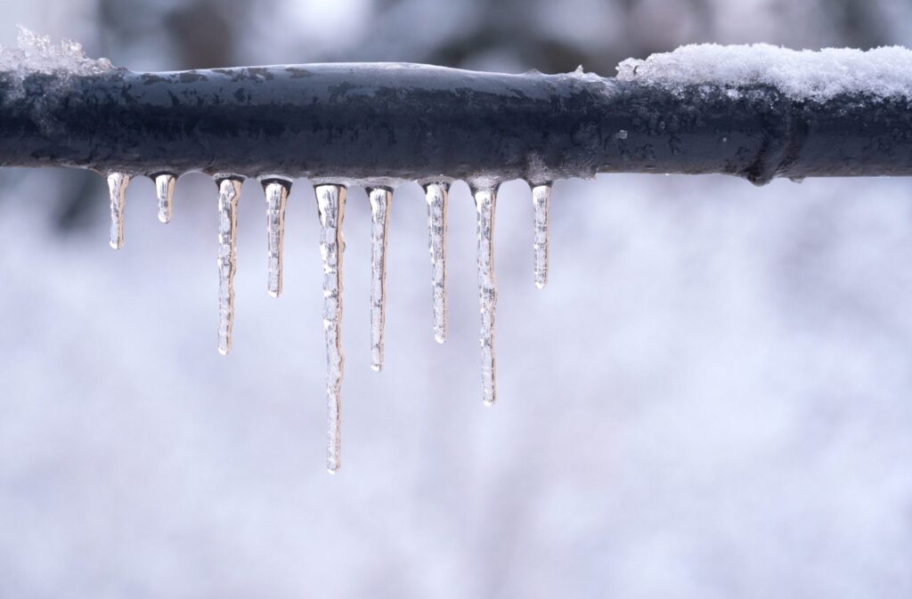 Several icicles hang from a snow-covered black metal pipe against a blurry white winter background.