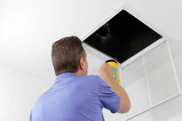 A man in a blue shirt uses a flashlight to inspect an open ceiling vent in a white room.