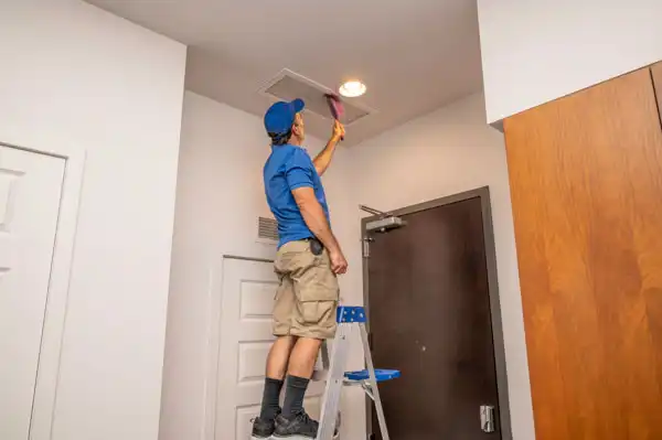 A person standing on a step ladder cleans an air vent on the ceiling with a brush in a residential interior.