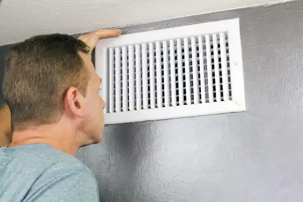 A person closely inspecting a white air vent mounted on a gray wall.