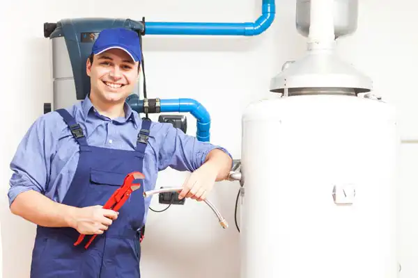 A plumber in blue overalls and cap holds tools while standing next to a water heater and pipes, smiling at the camera.