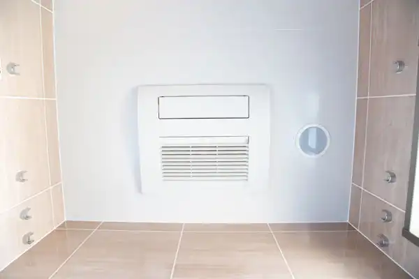 White wall-mounted bathroom ventilation fan with a vent and a round opening, surrounded by beige tiled walls and floor.
