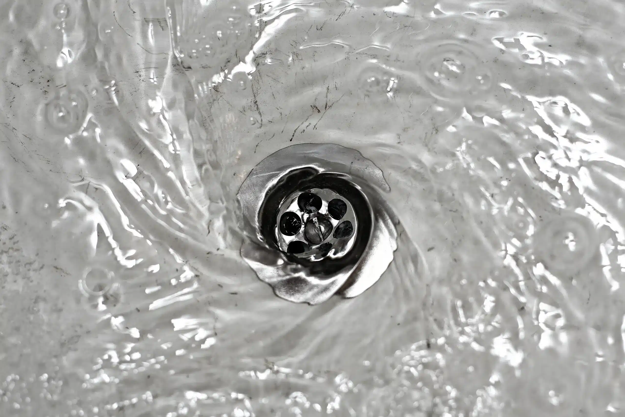 Water swirling and draining through a silver metal sink drain, creating ripples and small bubbles on the water surface.