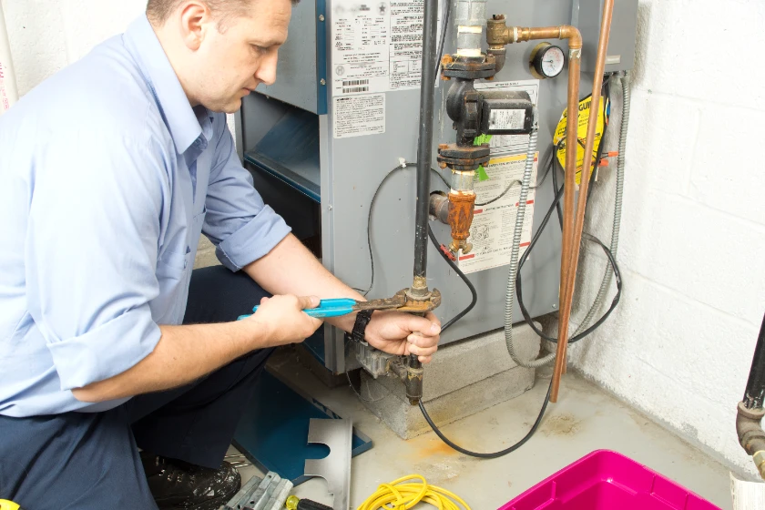 A man in a blue shirt uses a wrench to repair a pipe on a basement heating system, with tools and a pink container nearby.