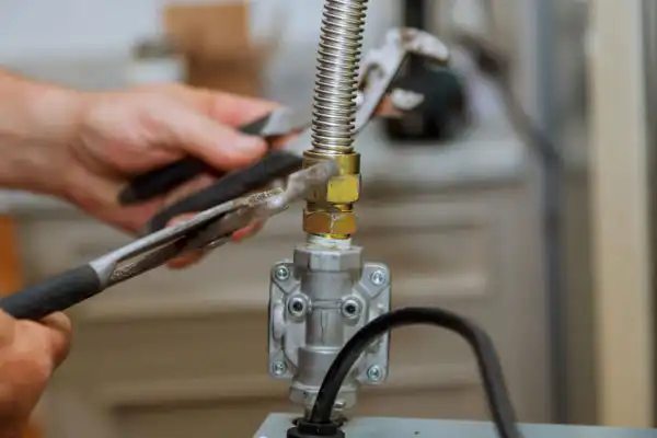 Hands using pliers to tighten a flexible metal gas hose connection on a valve in a home setting.