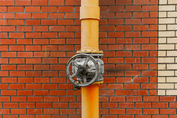 A yellow industrial pipe with a large metal valve wheel is mounted against a red and beige brick wall.