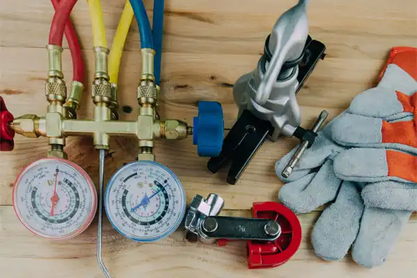 Assorted HVAC tools on a wooden surface, including manifold gauges with hoses, a pipe cutter, tubing bender, and a pair of work gloves.