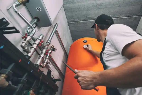 A worker in a cap uses a tool to adjust a valve on a large orange water heater in a utility room with exposed pipes.