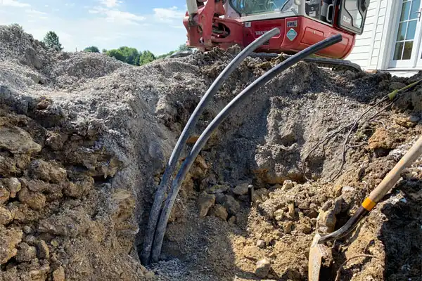 Three black utility cables emerge from underground in a dirt trench beside a house, with construction equipment and a shovel nearby.