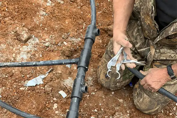 A person wearing camouflage clothing uses a tool to connect black irrigation pipes on bare soil.