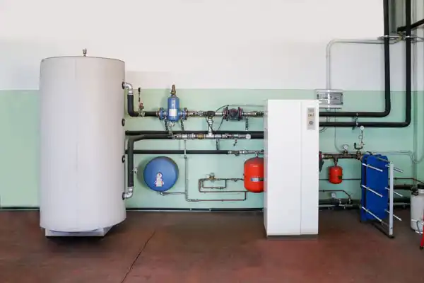 A modern heating system setup with a large white water tank, various pipes, gauges, a white control unit, red expansion tanks, and a blue heat exchanger on a clean indoor wall.