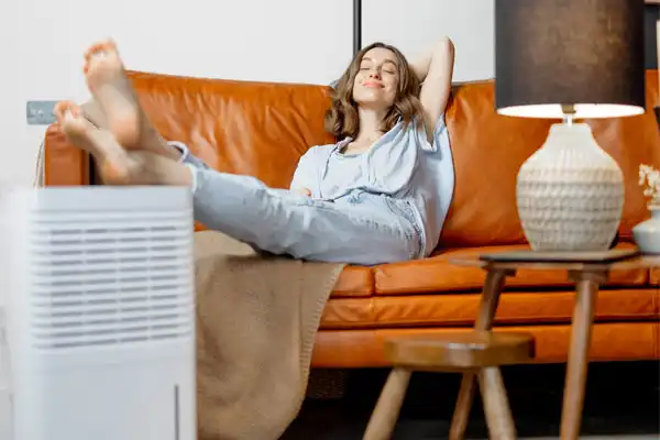 A woman relaxes on a brown leather sofa with her feet up, eyes closed, next to a dehumidifier and a table lamp.