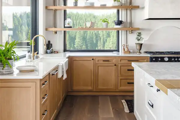 Modern kitchen with light wood cabinets, white countertops, open shelves with plants and dishes, large window overlooking a forested landscape, and a sink with a gold faucet.