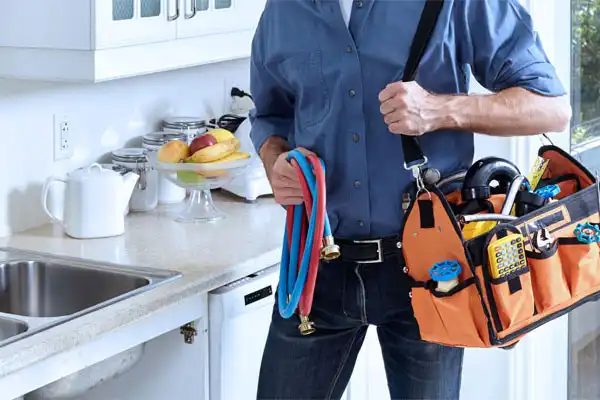 A person in a blue shirt stands in a kitchen holding plumbing hoses and an orange tool bag filled with various tools.