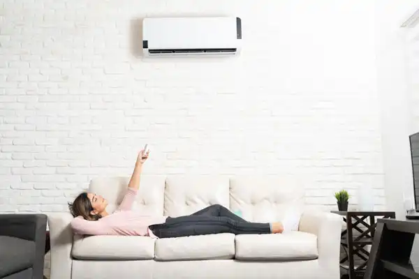 A person lying on a sofa points a remote control at a wall-mounted air conditioner in a bright living room.