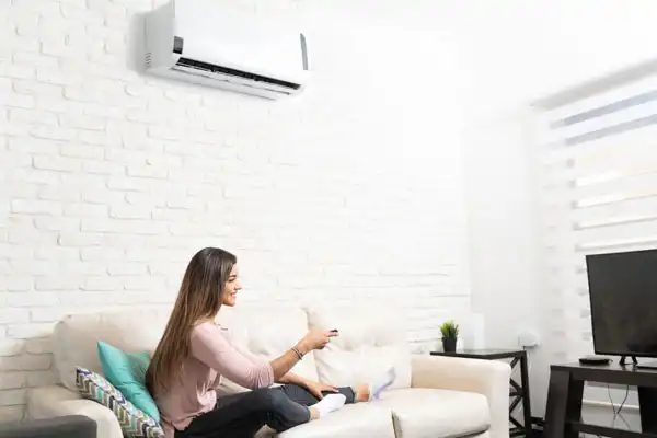 A woman sits on a couch holding a remote control, facing a wall-mounted air conditioner in a modern living room.