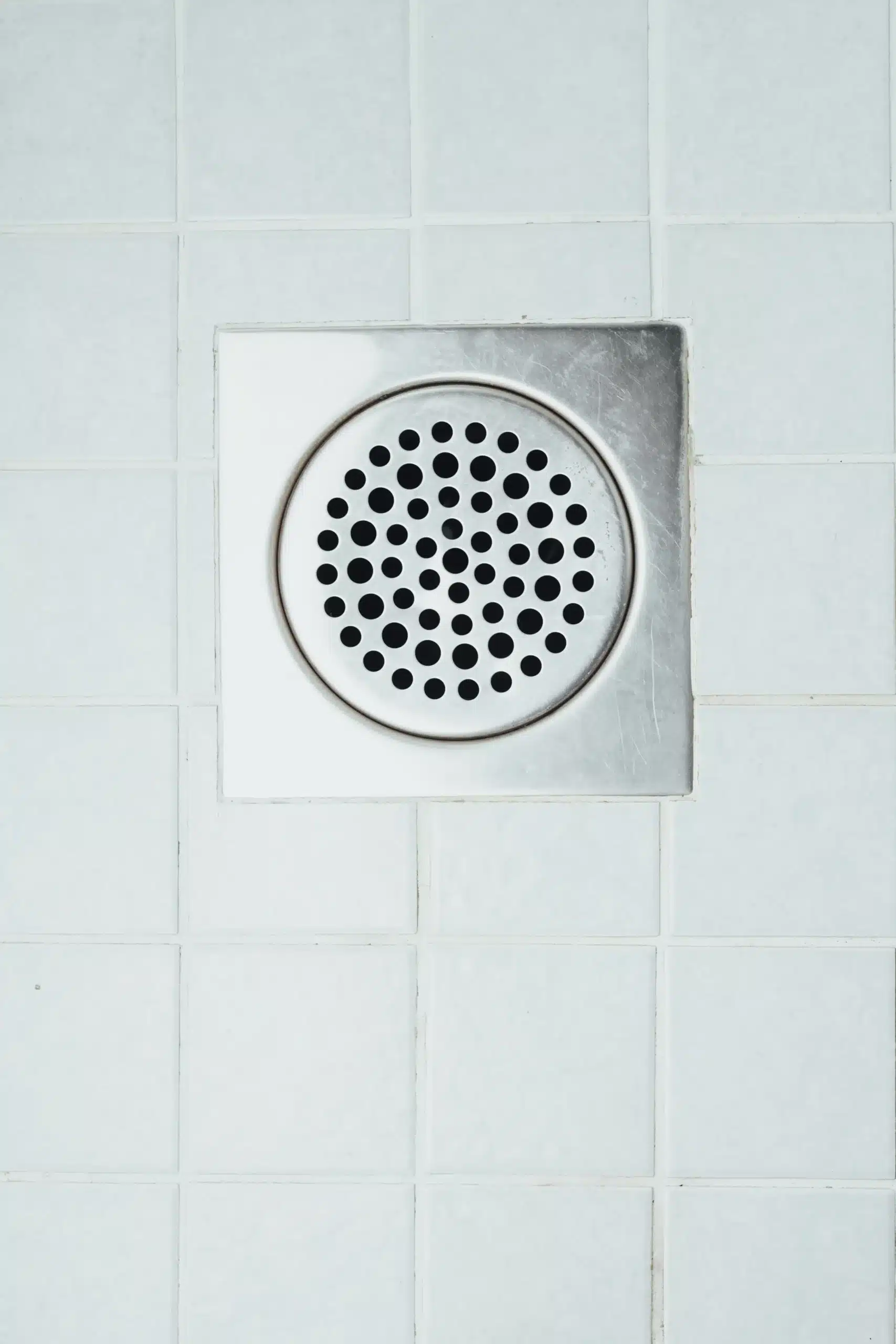 A metal drain with circular holes set in white tiled flooring.