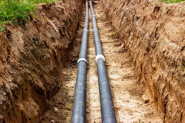 Two black pipes are laid parallel in a long, narrow trench dug in the dirt, with grass visible at the edges of the trench.