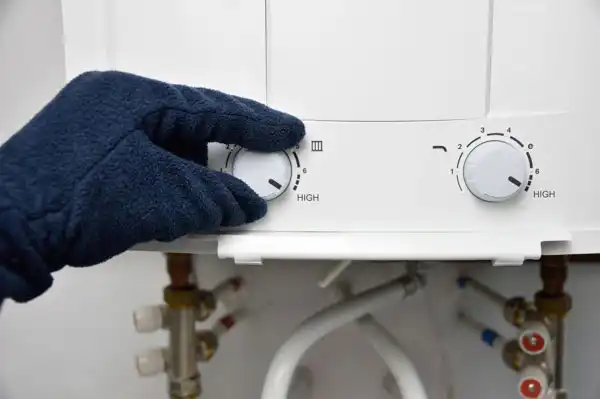 A gloved hand adjusts the temperature dial on a white water heater, with pipes and control knobs visible below.