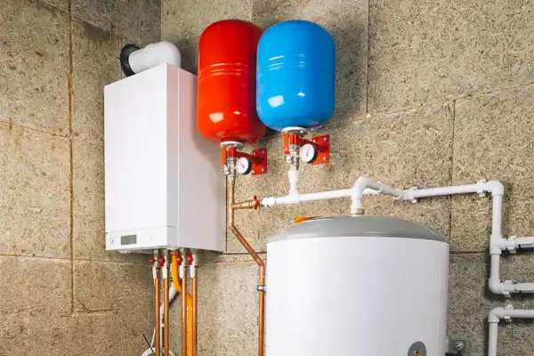 A wall-mounted boiler unit with red and blue expansion tanks is installed next to a hot water storage tank and white plumbing pipes in a utility room.
