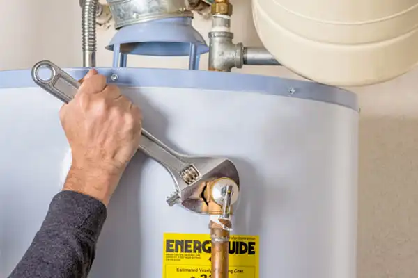 A person uses a large adjustable wrench to tighten a pipe fitting on top of a water heater, with an Energy Guide label visible on the unit.