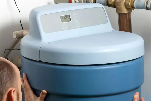 A person adjusts a large blue water softener with a digital control panel, installed near copper pipes.