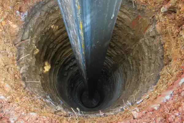 A large black pipe is being inserted vertically into a deep, circular hole in the ground surrounded by dirt.