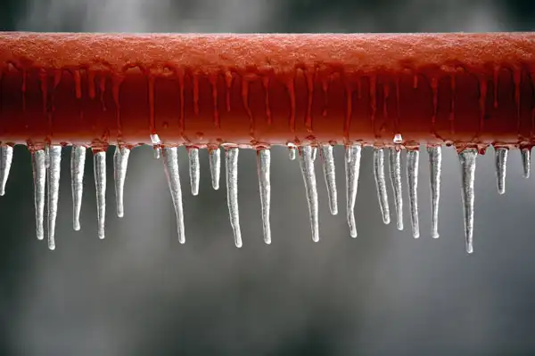 A row of icicles hangs from a red horizontal pipe, with ice droplets visible on the surface and a blurred gray background.