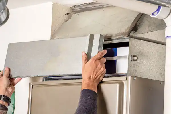 A person removes a metal panel from a furnace to access and replace the air filter inside the unit.