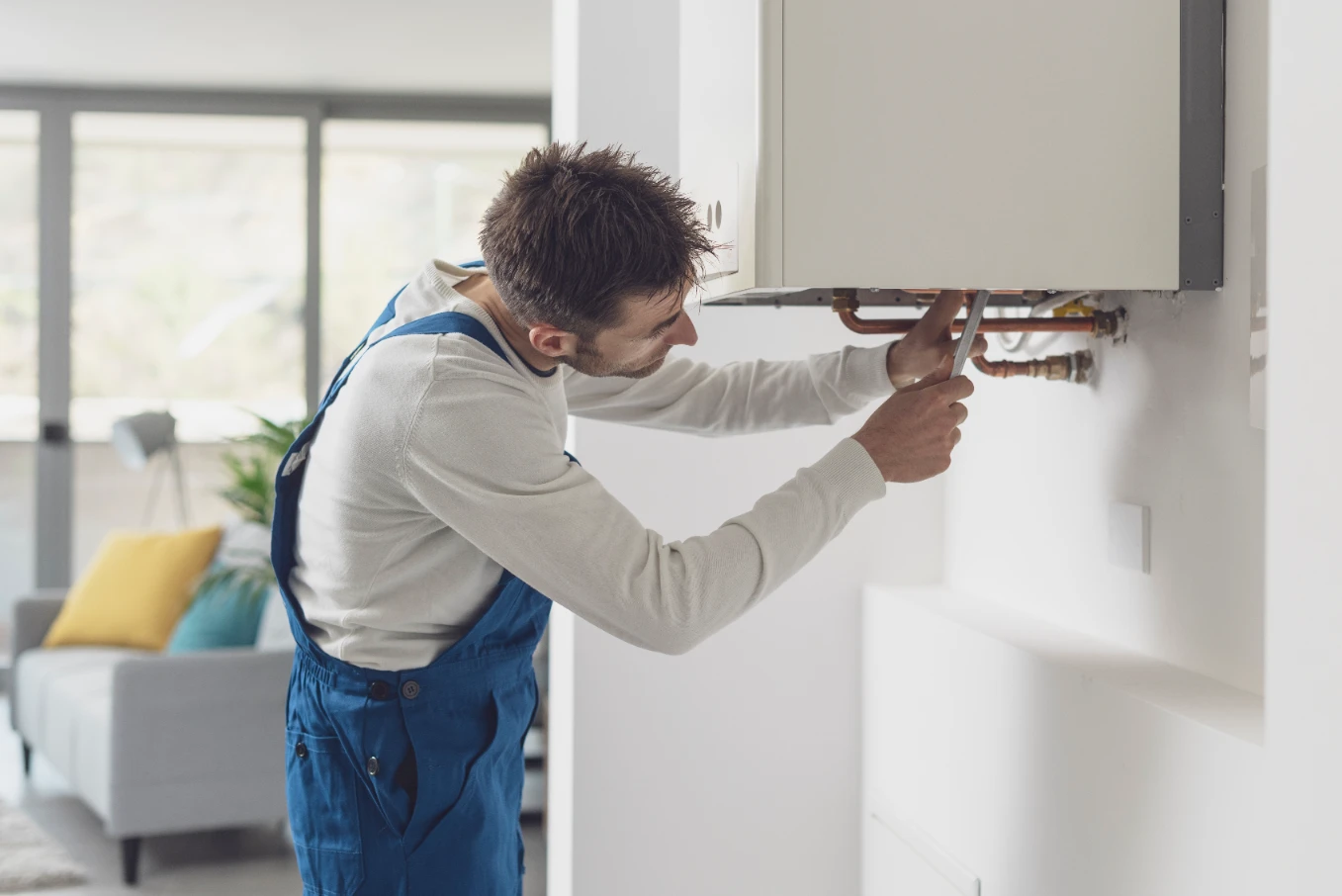 A man in blue overalls fixing or inspecting a wall-mounted boiler in a modern home.