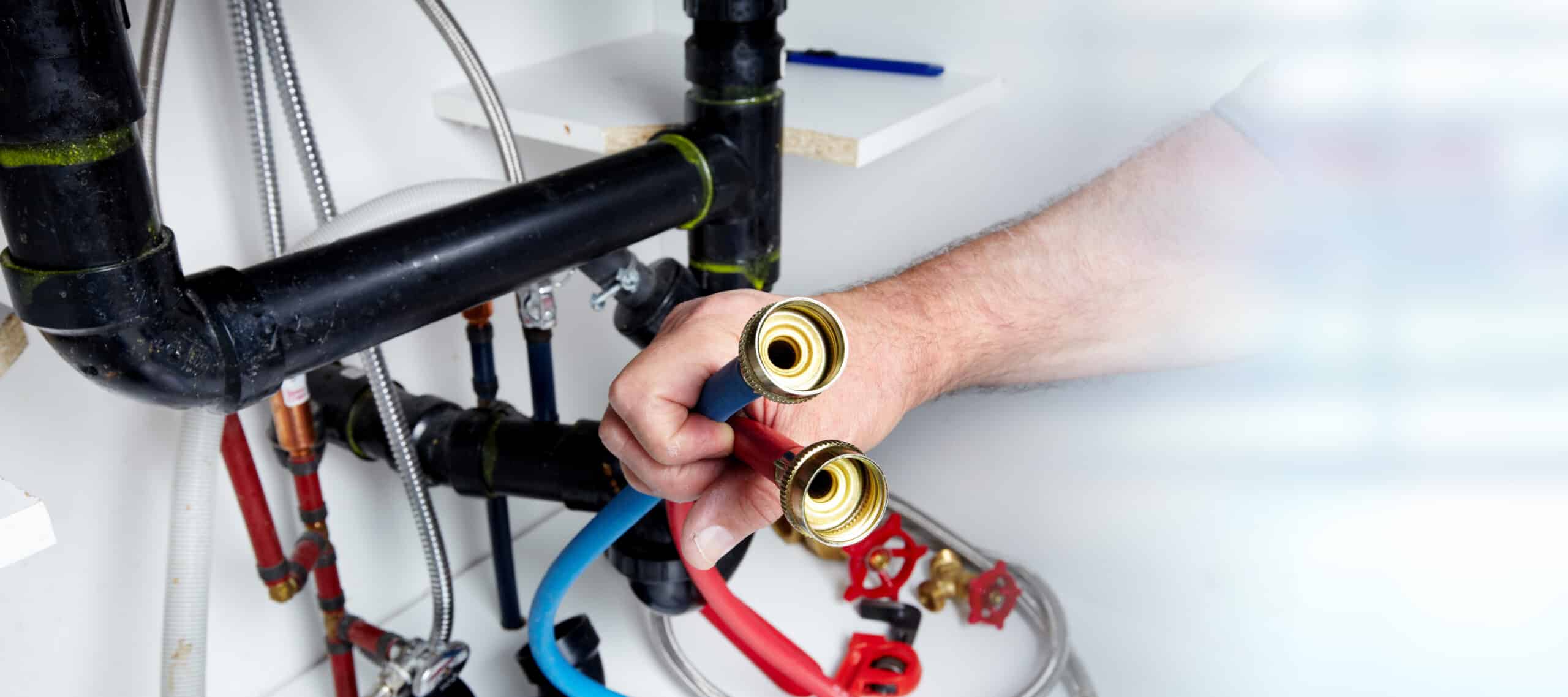 A person holds two hose connectors in front of exposed plumbing pipes beneath a sink.