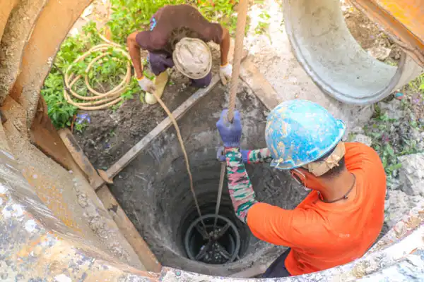 Two construction workers wearing helmets lower equipment into a deep concrete well using ropes, seen from above.